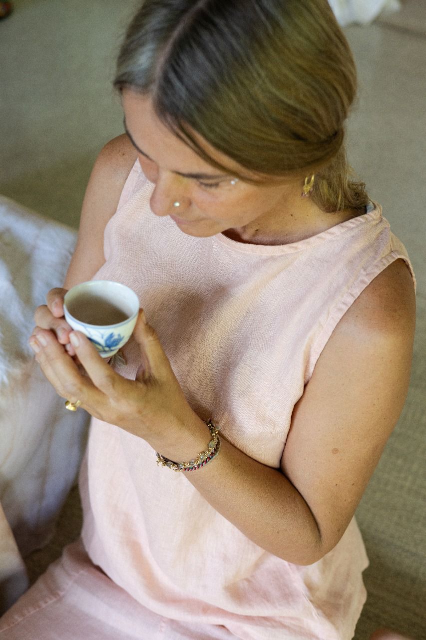 A person wearing a sleeveless crew-neck top in a warm rose color, sitting on the floor, holding a cup of tea, photographed from above.