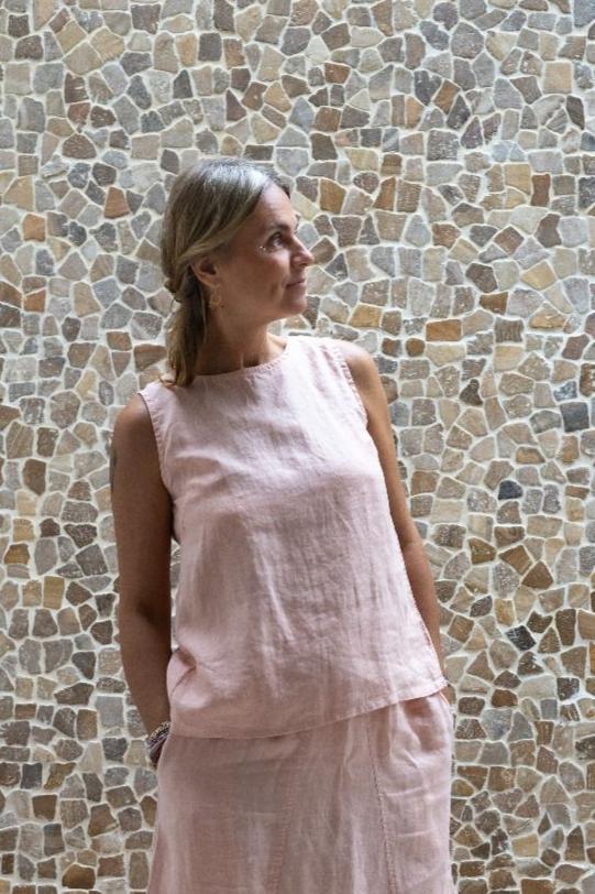 A person wearing a sleeveless crew-neck top in a warm rose color, with an open-back design from Shivani Divine Wear, standing against a textured wall.