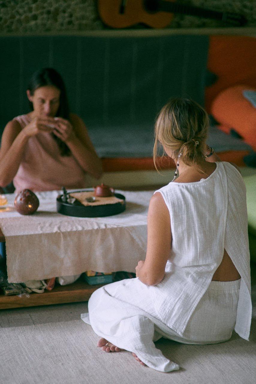 Two women wearing a sleeveless top and a matching skirt sitting on the floor, front view and drinking tea.