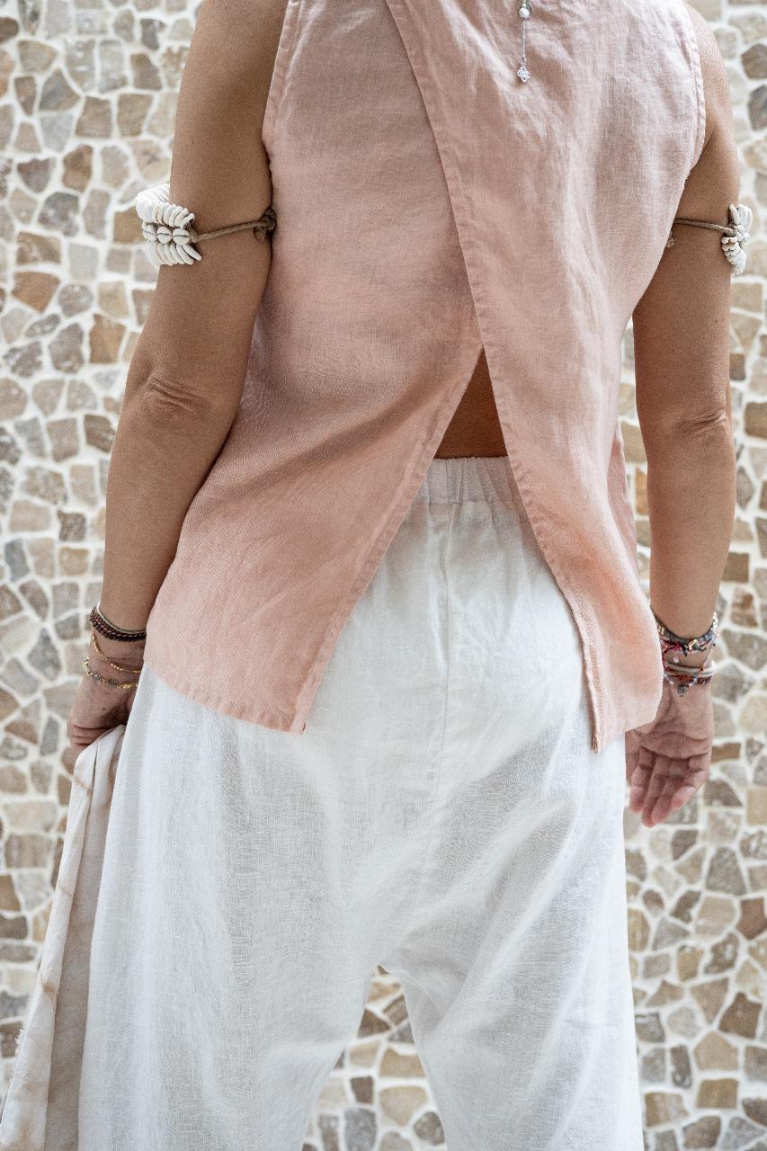 A woman standing against a stone wall backdrop, wearing white linen hipster pants and a matching rose color linen top, back view