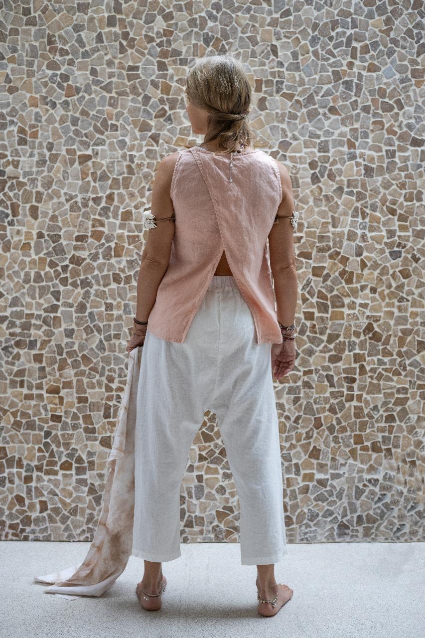 A woman standing against a stone wall backdrop, wearing white linen hipster pants and a matching rose color linen top, back view. All Shivani Divine Wear.
