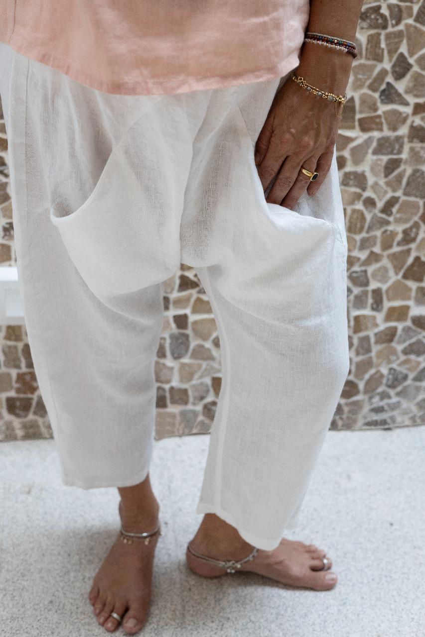A woman standing against a stone wall backdrop, wearing white linen hipster pants and a rose colored Top. Detail view