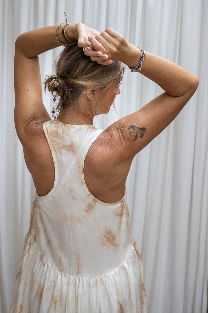 A person wearing a sleeveless, asymmetric hemline silk dress in front of a white background. Back view, detail.