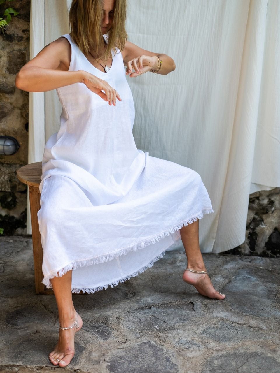 A woman wearing a white maxi v-neck linen dress, sitting on a chair in front of a white backdrop.