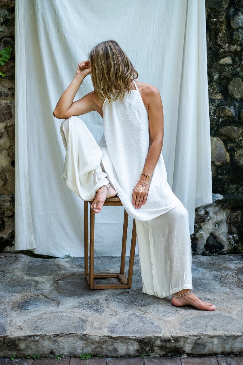 A woman sitting on a stool in front of a fabric white backdrop, modeling in silk white pants and white A line cotton dress.