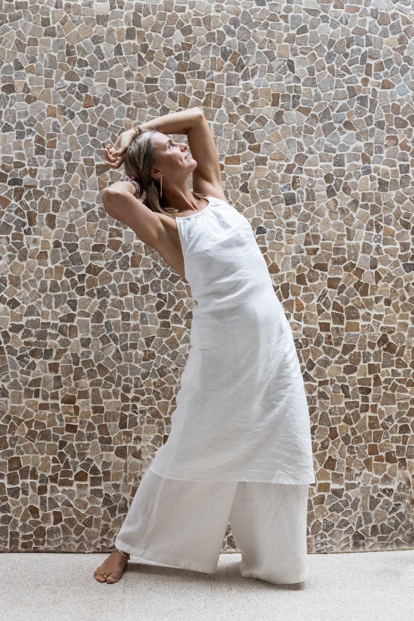 A woman modeling flowy white linen pants with a matching midi linen dress, standing against a backdrop with a stone wall.