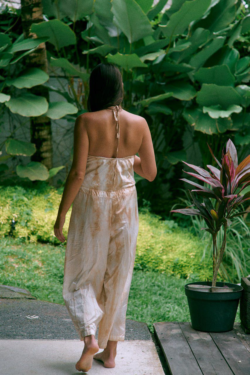 A woman wearing a beige tea-dye elegant silk romper with a halter-style neckline and wide-leg silhouette, standing with greenery in the background. Back view