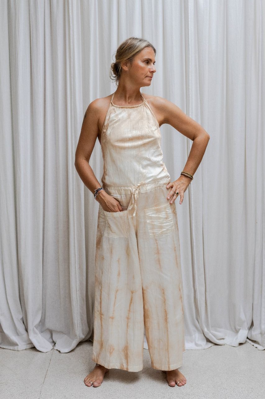 A woman wearing a beige tea-dye elegant silk romper with a halter-style neckline and wide-leg silhouette, standing with a white backdrop in the background.