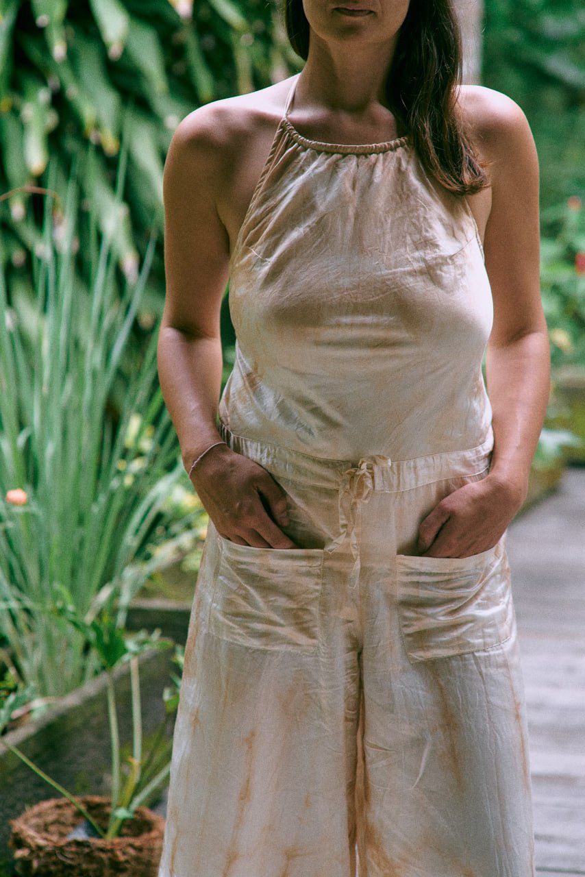 A woman wearing a beige tea-dye elegant silk romper with a halter-style neckline and wide-leg silhouette, standing on a wooden surface with greenery in the background. Close up view.