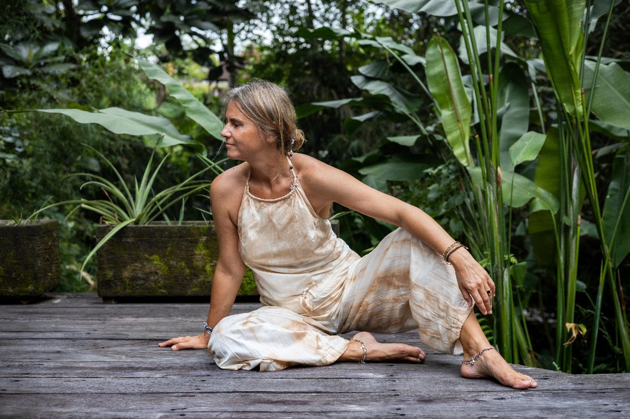 A woman wearing a beige tea-dye elegant silk romper with a halter-style neckline and wide-leg silhouette, sitting on a wooden surface with greenery in the background.