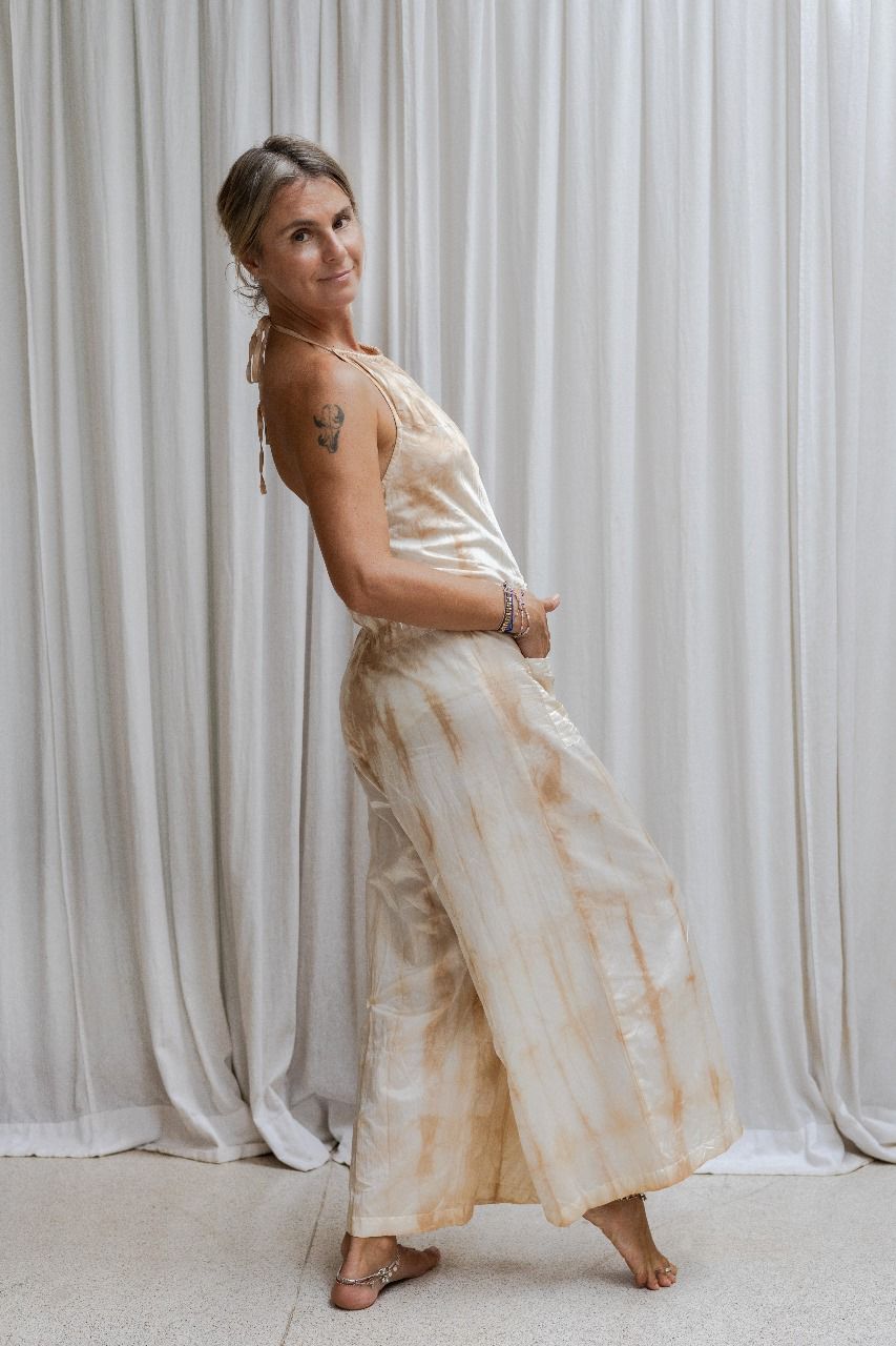 A woman wearing a beige tea-dye elegant silk romper with a halter-style neckline and wide-leg silhouette, standing with a white backdrop in the background. Side view 2