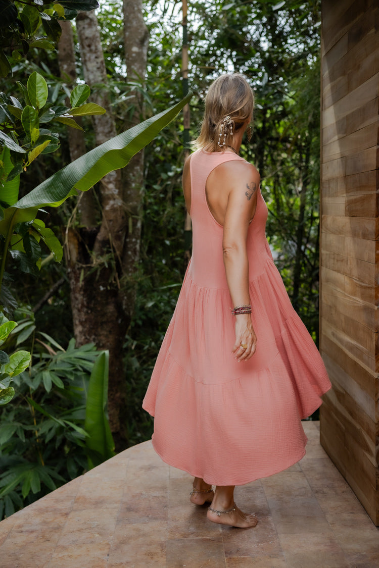 A woman from back, in a rose color asymmetric cotton dress with a sleeveless design standing near a wooden structure and lush greenery in the background.