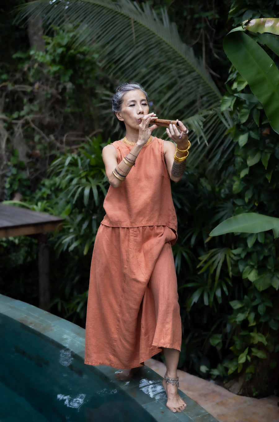 A woman walking along the edge of a swimming pool and wearing a sleeveless pink top and a matching skirt made by Shivani Divine Wear and in front of a lush green background.