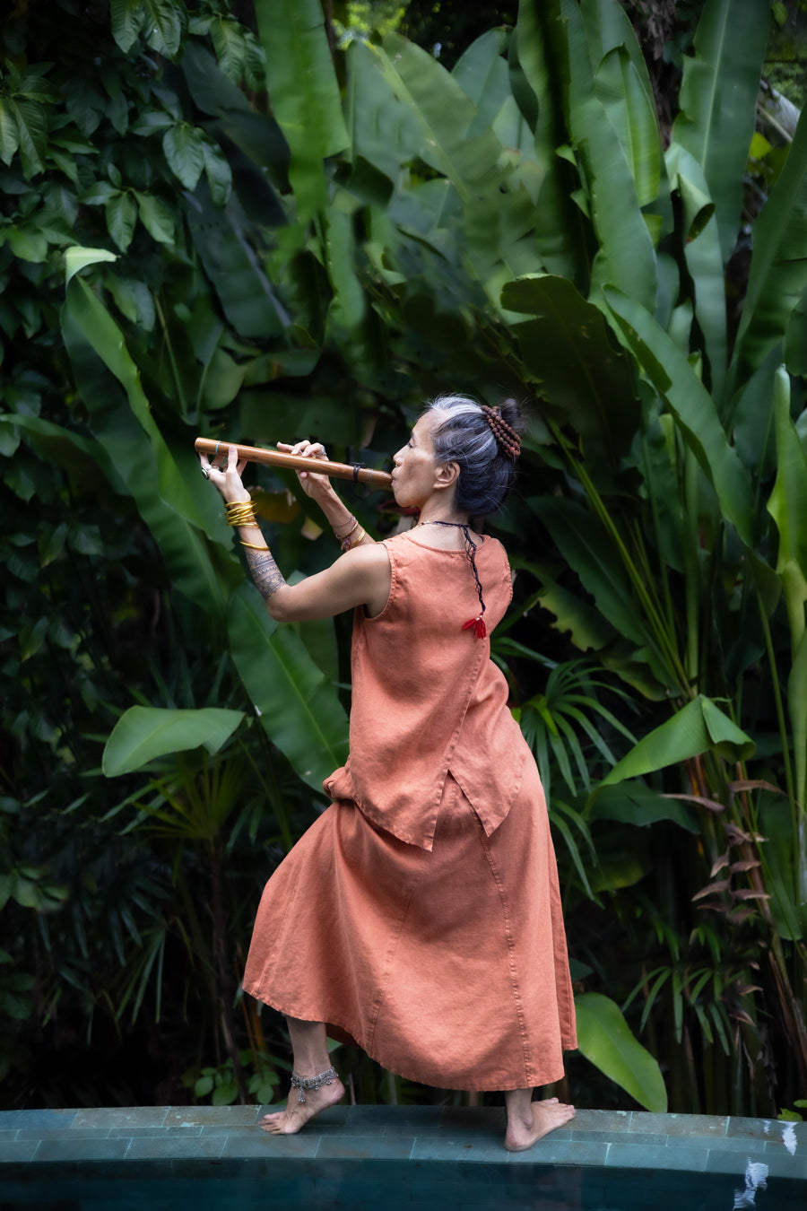 A woman wearing a sleeveless light terracotta top and a matching skirt standing in front of a lush green jungle background and playing a flute. Back view.