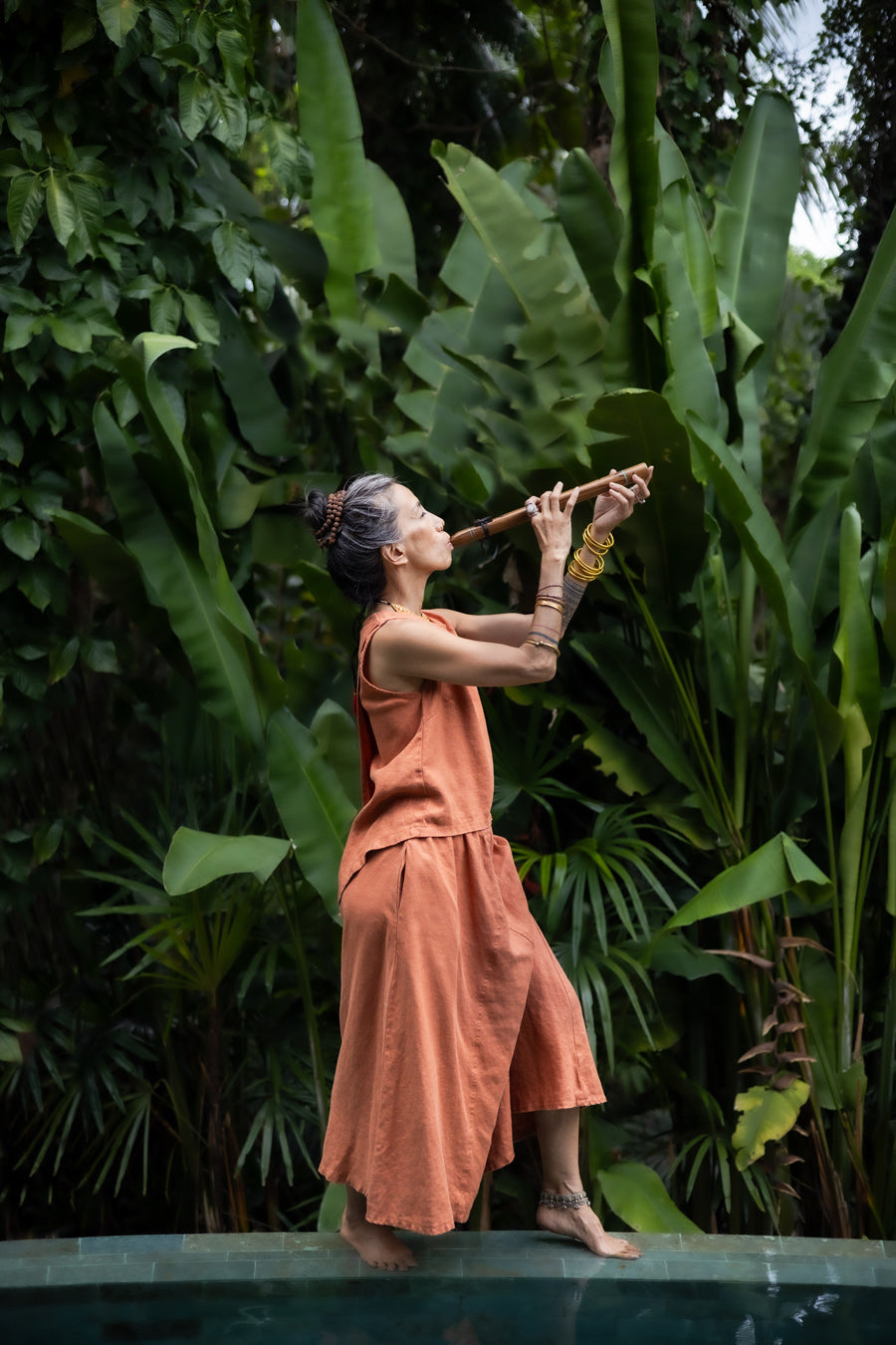 A woman wearing a sleeveless light terracotta top and a matching skirt standing in front of a lush green jungle background and playing a flute. Side view.