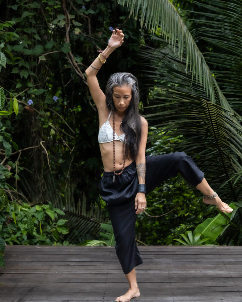A woman standing against a lush green backdrop, wearing black linen hipster pants and a white cotton bra. All Shivani Divine Wear.