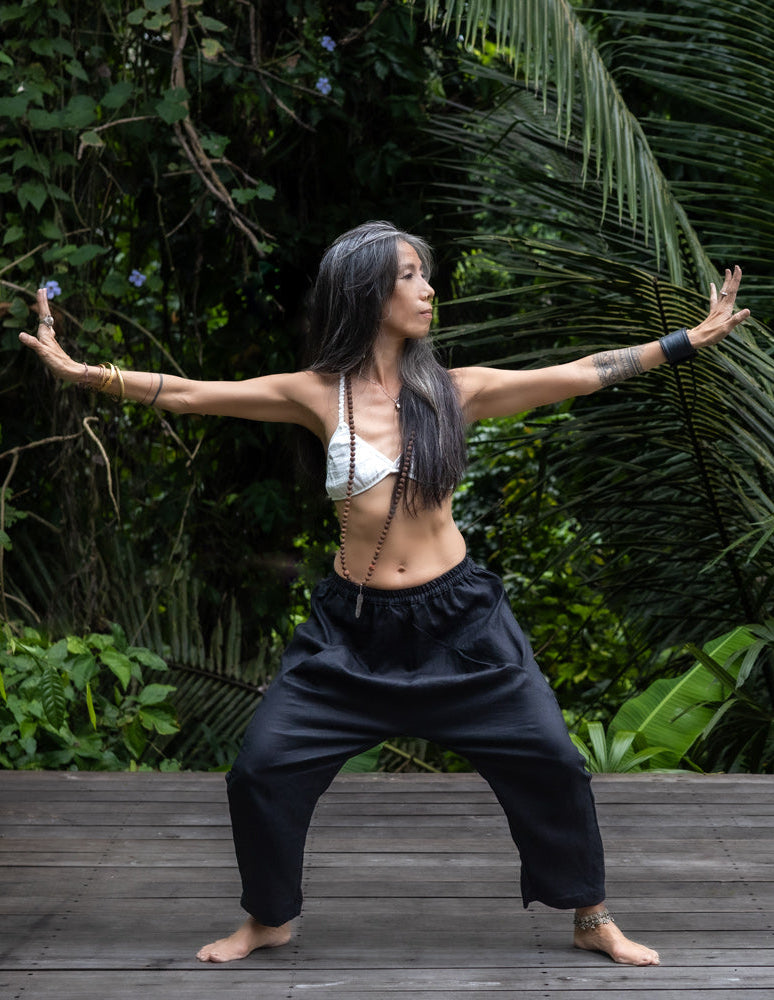 A woman standing against a lush green backdrop, wearing black linen hipster pants and a white cotton bra, front view. All Shivani Divine Wear.