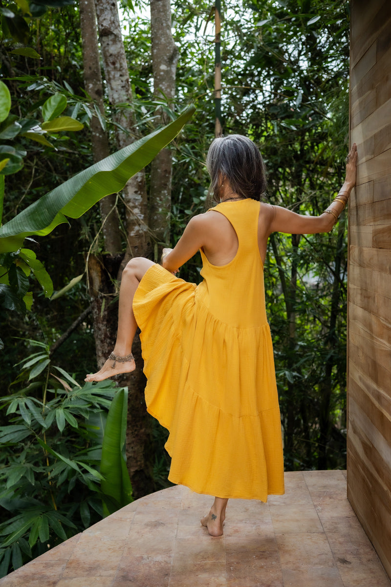 A woman from the back in a yellow asymmetric cotton dress with a sleeveless design standing with her one leg raised, near a wooden structure and lush greenery in the background.