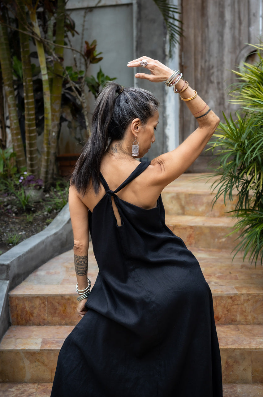 A woman standing on outdoor steps, in a black flowing linen dress, with arms extended gracefully.