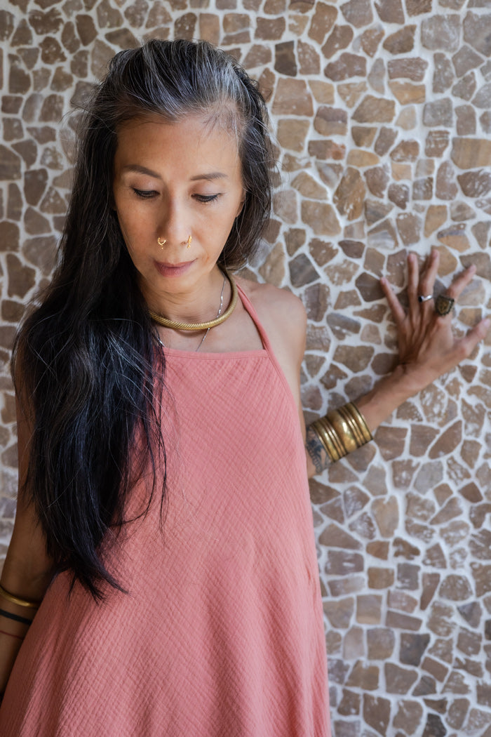 A woman wearing a rose color, open back tie cotton dress with a texture that drapes over the body.