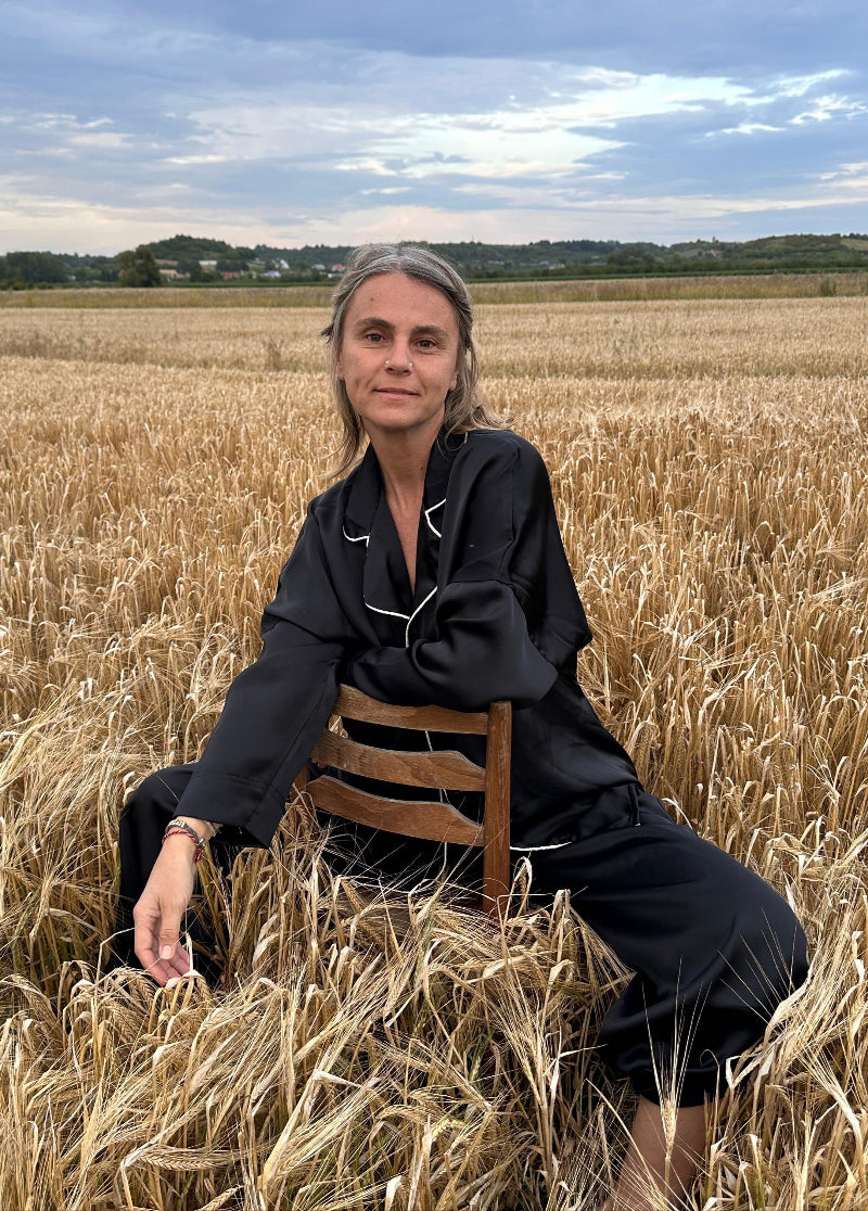 Woman sitting confidently on a wooden chair in the middle of a golden wheat field, wearing a black silk pajama set with white piping.