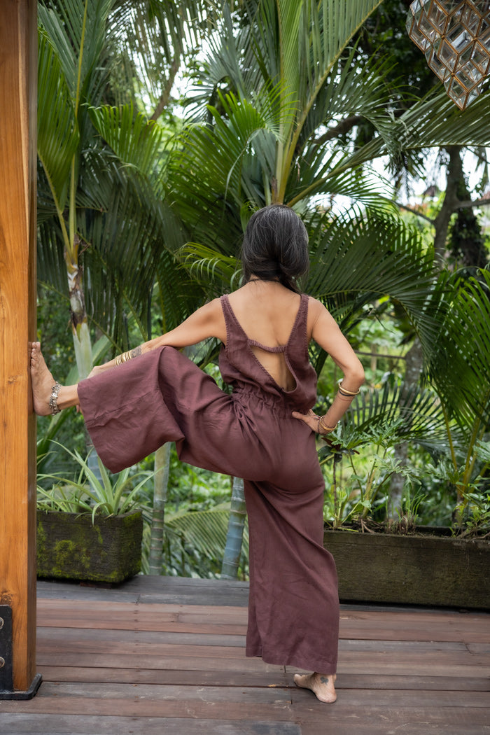 A woman, her back turned to the camera, wearing a brown linen romper with wide legs and a deep V-back, in a lush green landscape