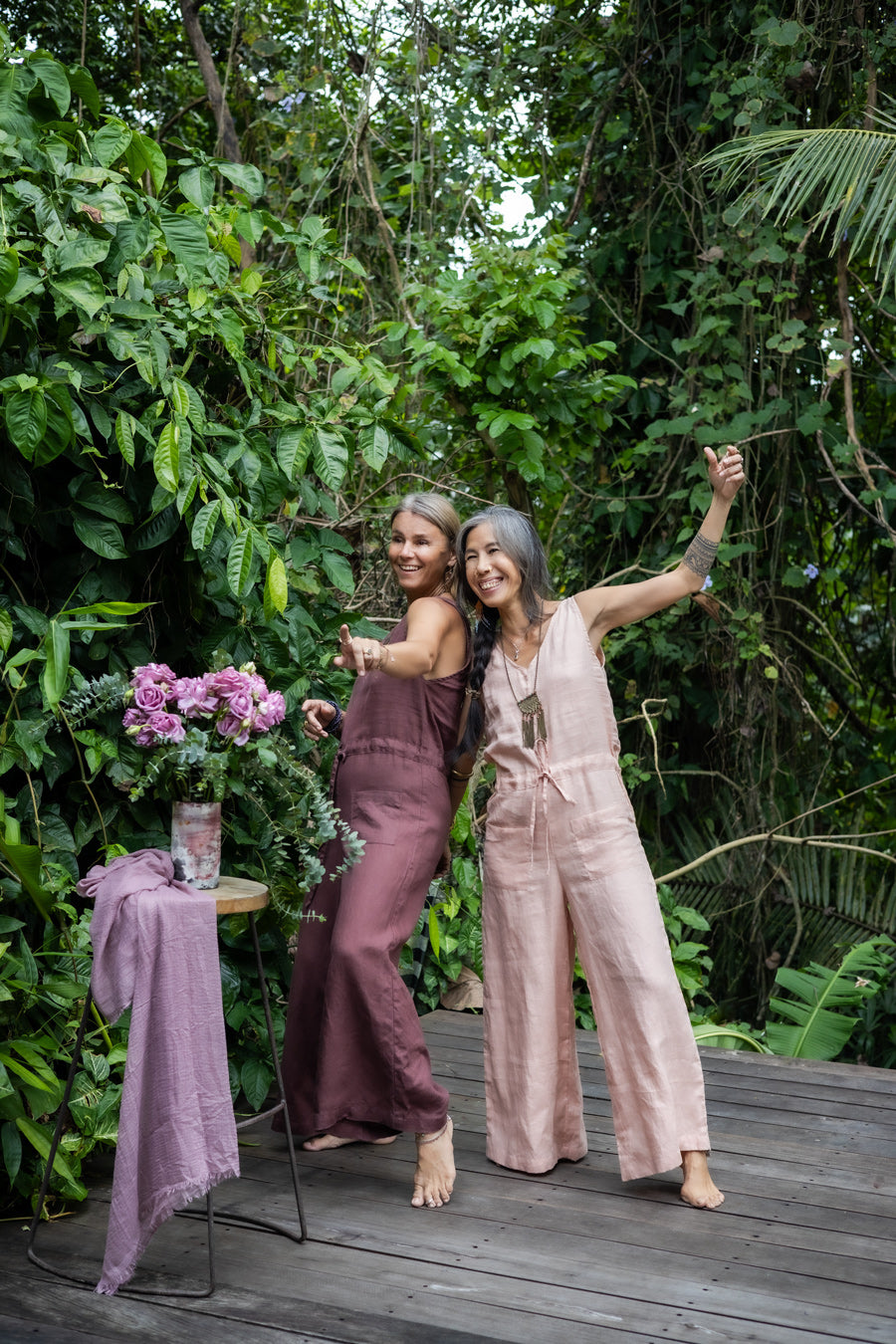 2 women wearing a rose pink and brown linen romper with wide legs and a V-neckline, standing in front of a lush green background.