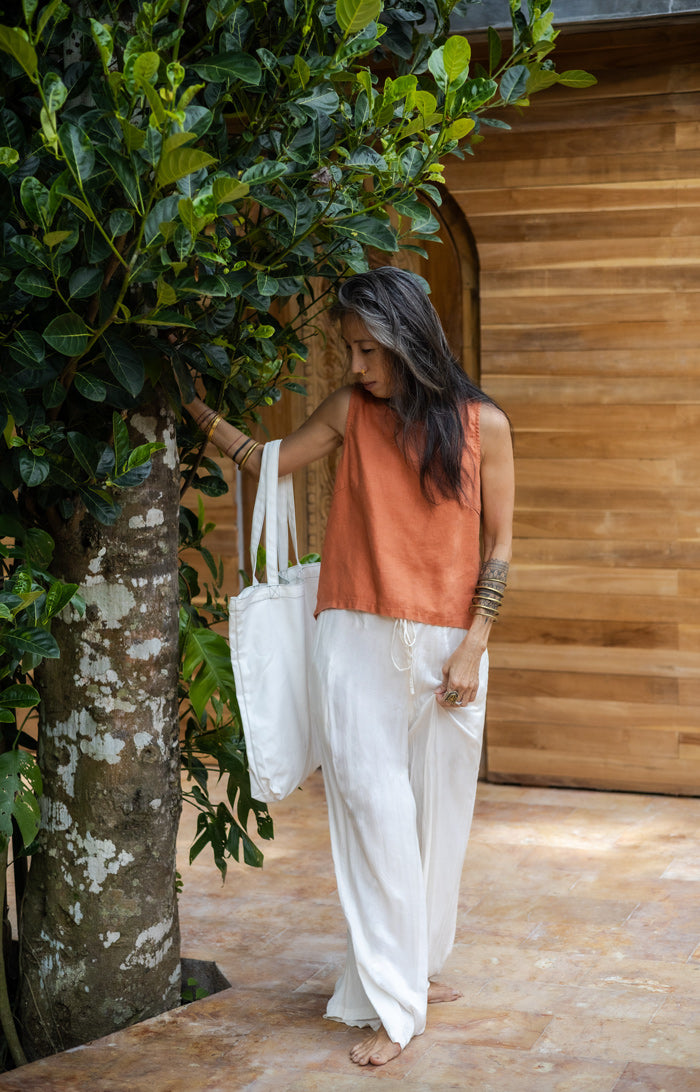 A woman standing, with long hair modeling silk white pants and a rose colored linen sleeveless top with a cotton tote bag from Shivani Divine Wear as well.