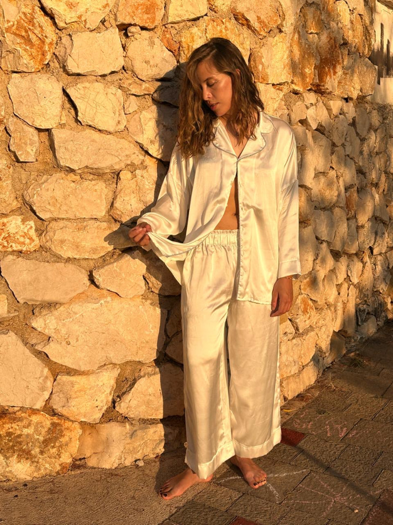Woman in a white silk pajama outfit standing against a stone wall.