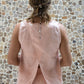 A person wearing a sleeveless crew-neck top in a warm rose color, with an open-back design, standing with their back towards the camera against a textured wall.