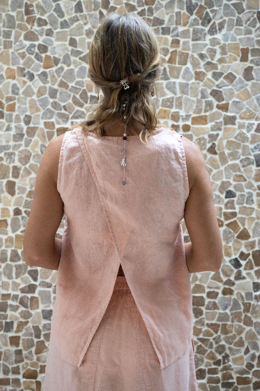 A person wearing a sleeveless crew-neck top in a warm rose color, with an open-back design, standing with their back towards the camera against a textured wall.