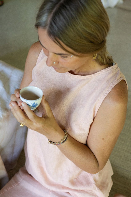 A person wearing a sleeveless crew-neck top in a warm rose color, sitting on the floor, holding a cup of tea, photographed from above.