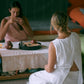 Two women wearing a sleeveless top and a matching skirt sitting on the floor, front view and drinking tea.