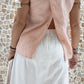 A woman standing against a stone wall backdrop, wearing white linen hipster pants and a matching rose color linen top, back view