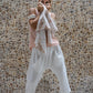 A woman standing against a stone wall backdrop, wearing white linen hipster pants and a matching rose color linen top, with her hands raised to her face. All Shivani Divine Wear.