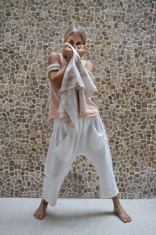 A woman standing against a stone wall backdrop, wearing white linen hipster pants and a matching rose color linen top, with her hands raised to her face. All Shivani Divine Wear.