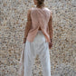 A woman standing against a stone wall backdrop, wearing white linen hipster pants and a matching rose color linen top, back view. All Shivani Divine Wear.
