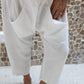 A woman standing against a stone wall backdrop, wearing white linen hipster pants and a rose colored Top. Detail view