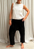 A woman standing against a white backdrop, wearing black linen hipster pants and sleeveless cotton top. Front view.