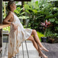 A person sitting on a chair wearing a sleeveless, asymmetric hemline silk dress in an outdoor setting with greenery in the background.