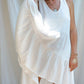 A woman in a white asymmetric cotton dress with a sleeveless design standing in front of a white fabric backdrop, holding her dress with one hand and smiling. 