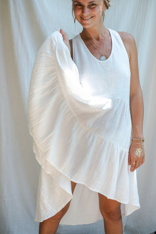 A woman in a white asymmetric cotton dress with a sleeveless design standing in front of a white fabric backdrop, holding her dress with one hand and smiling. 