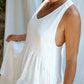 A woman in a white asymmetric cotton dress with a sleeveless design standing in front of a white backdrop, close up.