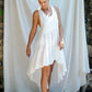 A woman in a white asymmetric cotton dress with a sleeveless design standing in front of a white fabric backdrop and stone wall.