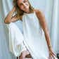 A woman wearing a white, open back tie cotton dress with a texture, sitting on a wooden stool. She is pairing the mini dress with white silk pants.