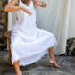 A woman wearing a white maxi v-neck linen dress, sitting on a chair in front of a white backdrop.