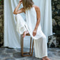 A woman sitting on a stool in front of a fabric white backdrop, modeling in silk white pants and white A line cotton dress.