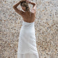 A woman modeling flowy white linen pants with a matching midi linen dress, standing against a backdrop with a stone wall. Back view.