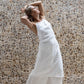 A woman modeling flowy white linen pants with a matching midi linen dress, standing against a backdrop with a stone wall.