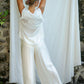 A woman standing her back to the camera in front of a fabric white backdrop, modeling in silk white pants and silk white relaxed top.